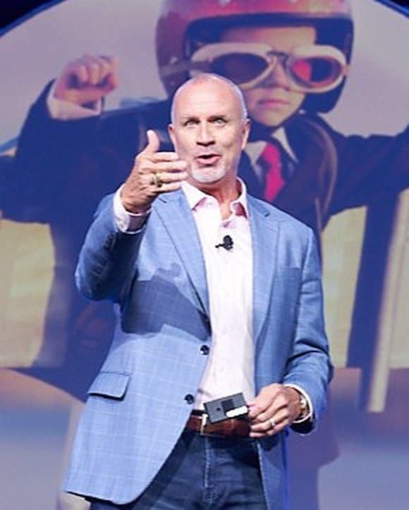 Dr. Randy Ross speaking on stage in a light blue blazer, gesturing toward the audience.